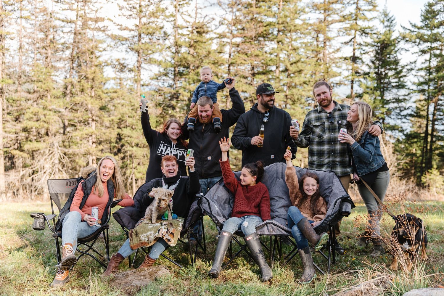 A group of people pose together with their dogs in a forest setting during autumn.