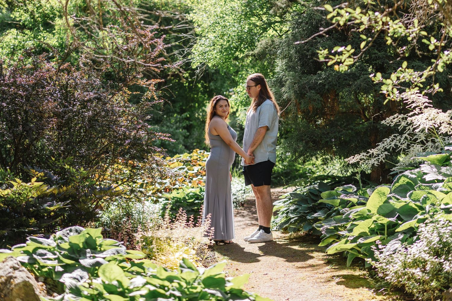 A scenic wooded garden pathway leads through lush green foliage and dappled sunlight on a summer day.