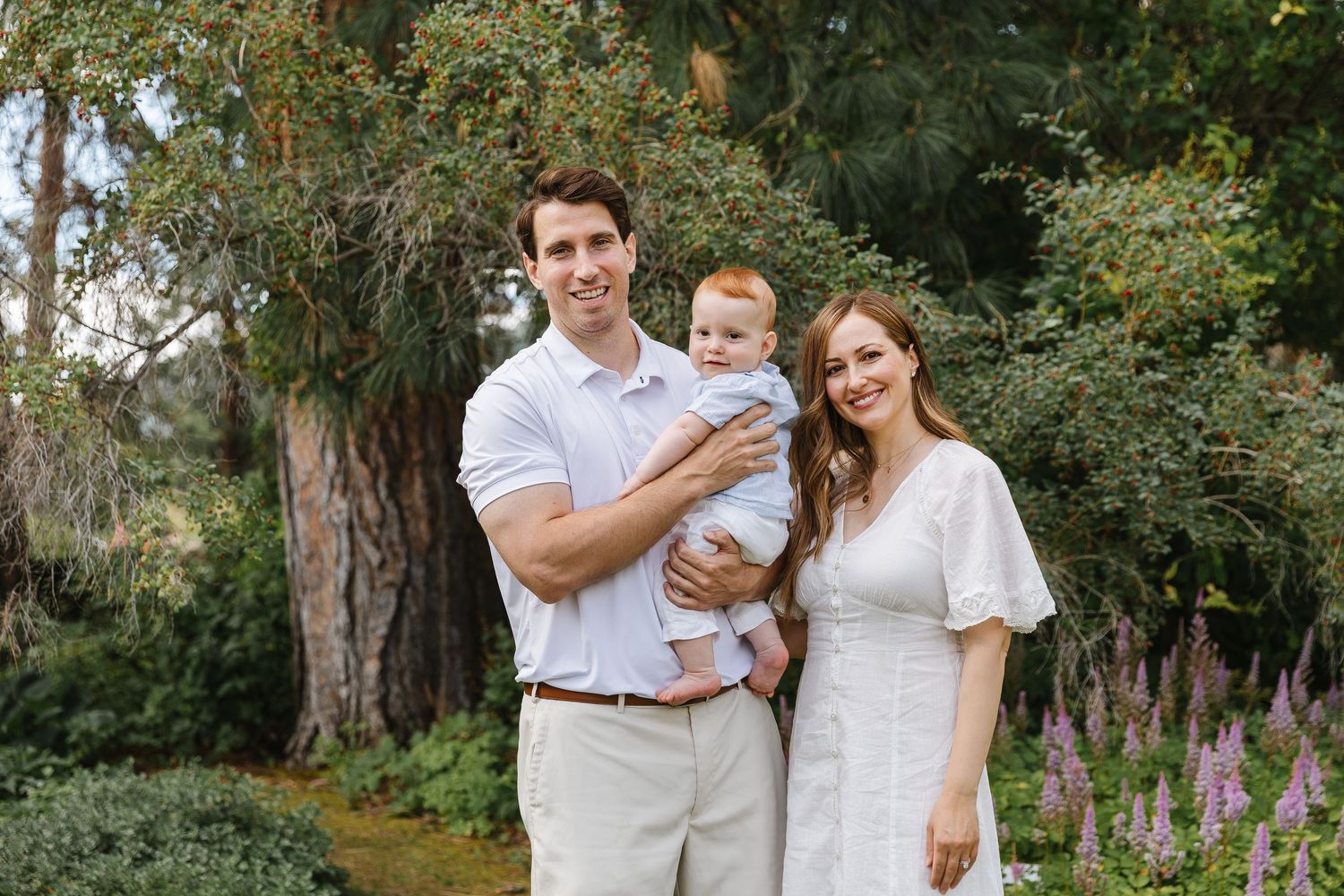 A family wearing coordinated white outfits poses together in a lush garden setting with blooming purple flowers.