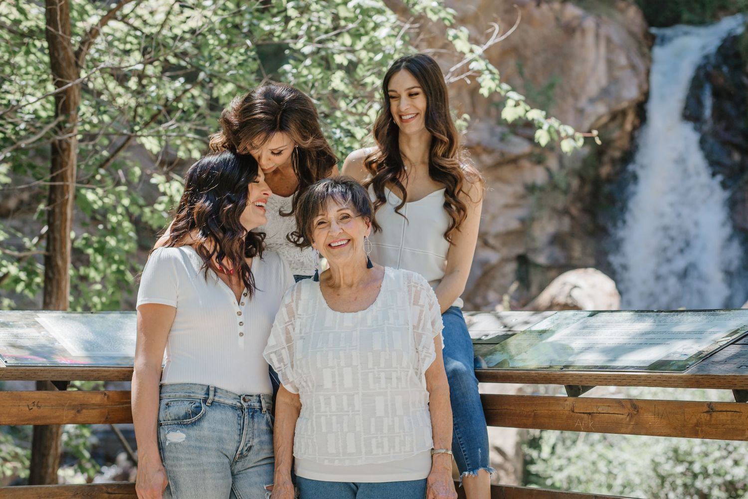 Three people in casual white tops pose together on a wooden deck with a scenic waterfall in the background.