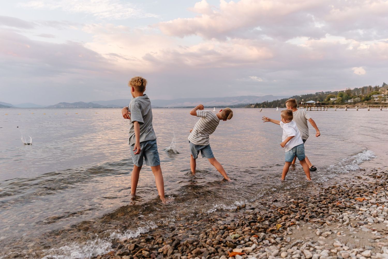 Three people skipping rocks on a pebble beach at sunset with mountains and coastal homes in the background.