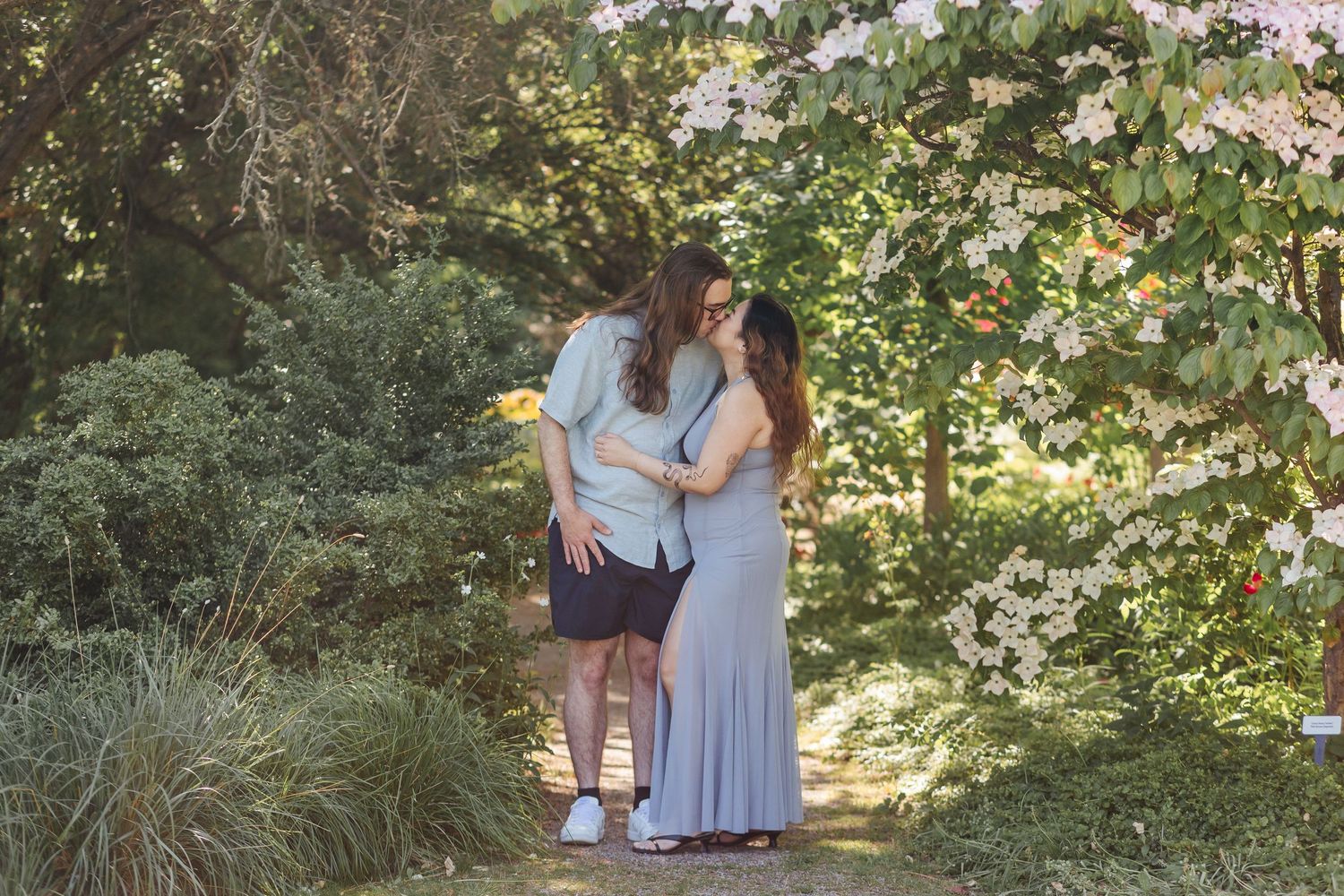A couple in light blue attire poses together in a lush garden setting during a sunny outdoor photoshoot.