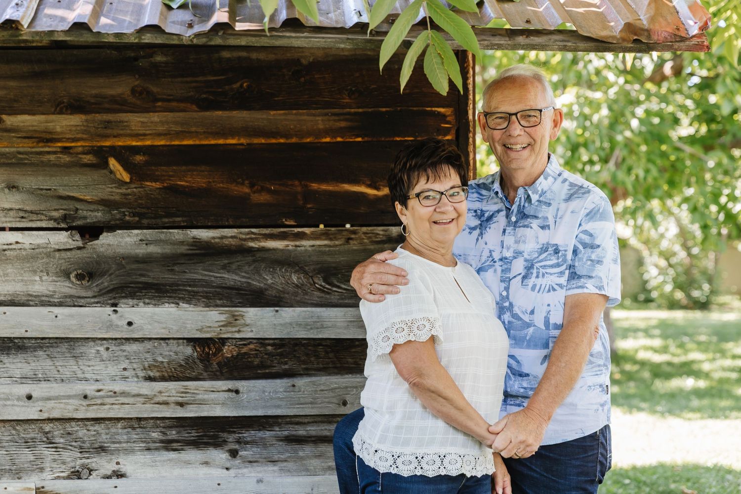 Couple in casual attire sitting and embracing on rustic wooden bench near log cabin in outdoor garden setting.