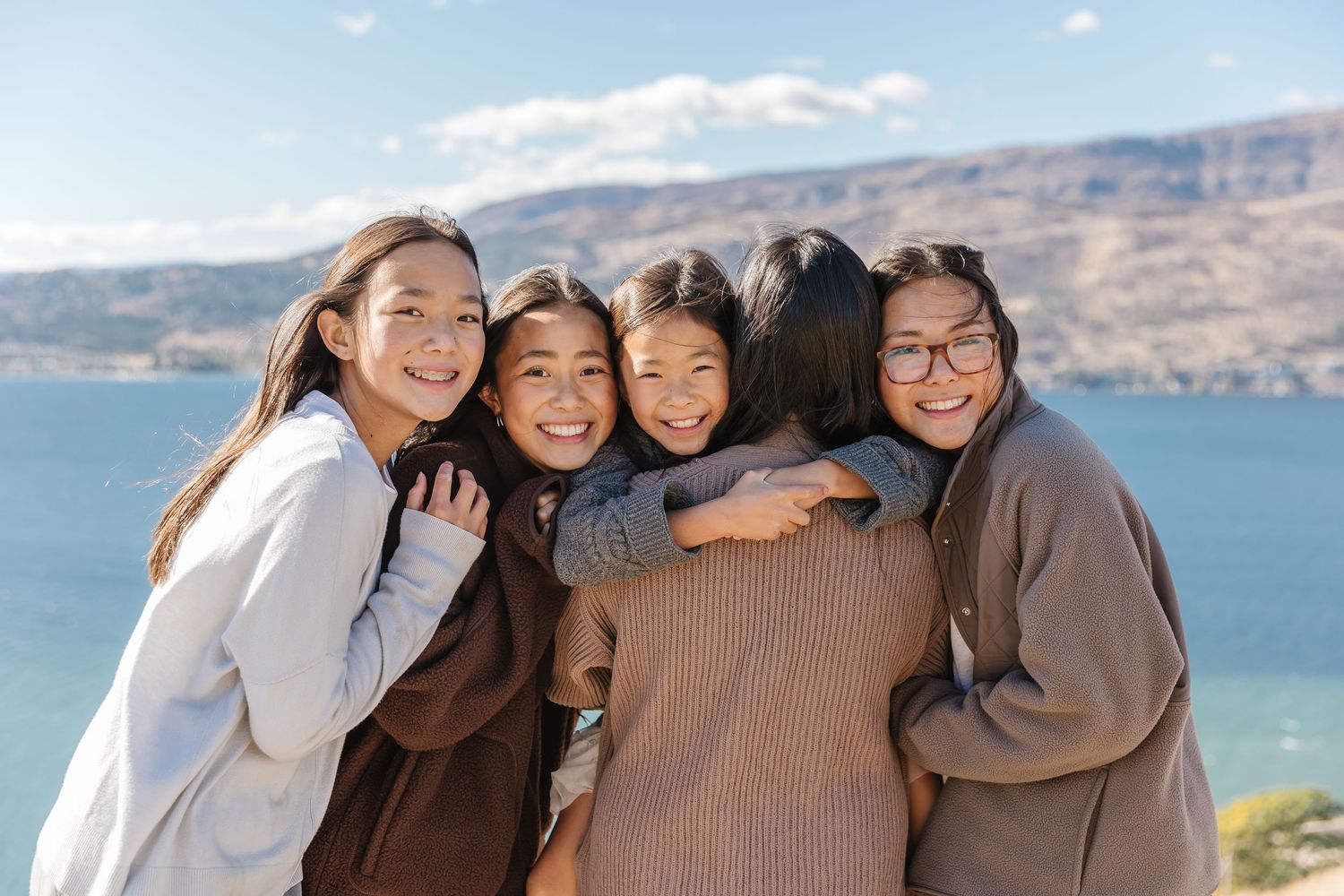 A group of friends embrace and smile together while posing outdoors against a scenic mountain and lake backdrop.