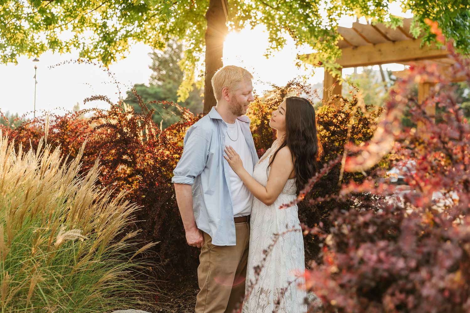A romantic couple embraces in a garden setting with golden sunlight filtering through autumn foliage and ornamental grasses.