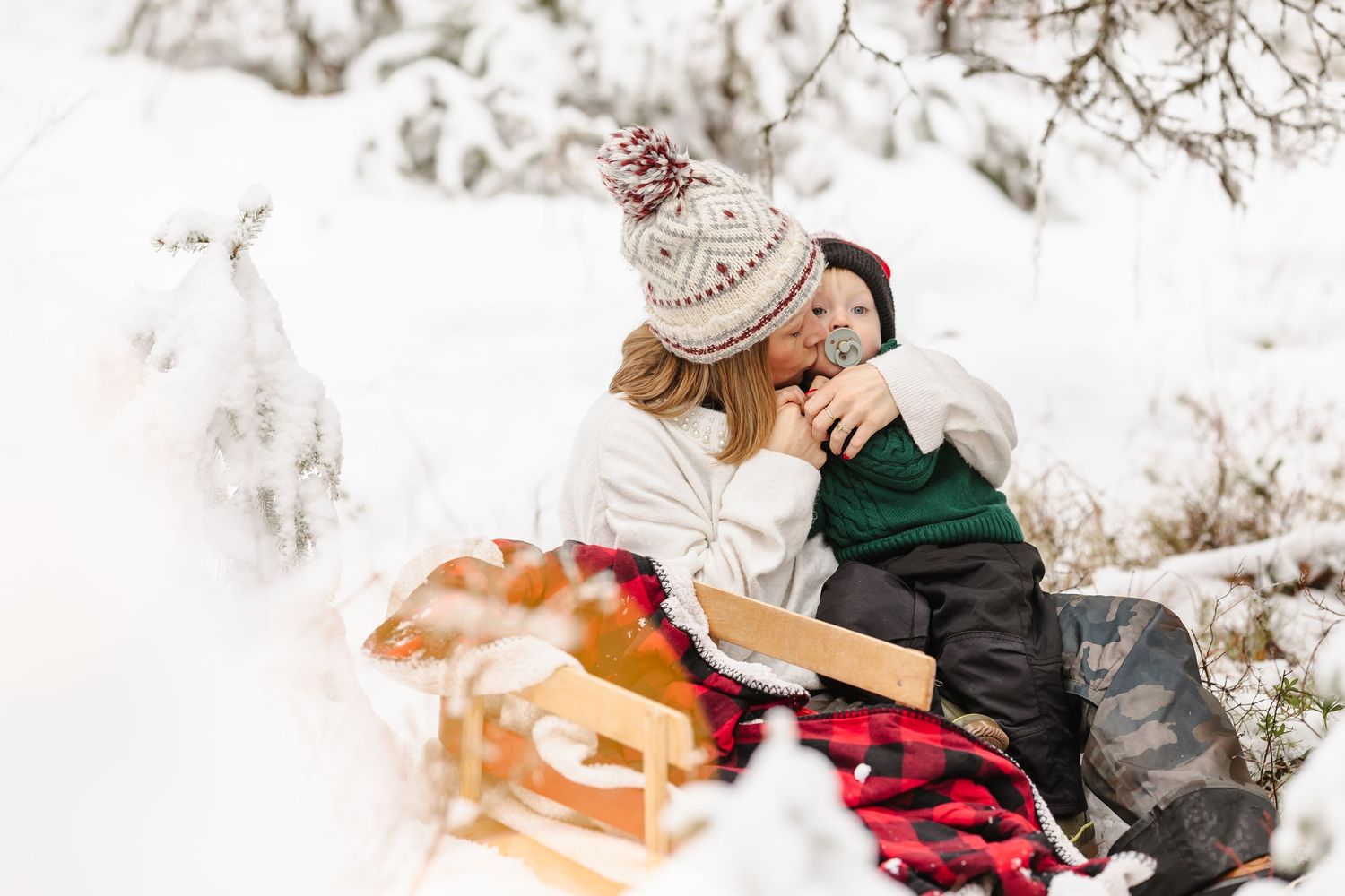 A person sits in deep snow during winter wearing a black coat and white knit hat with snowflakes falling around them.
