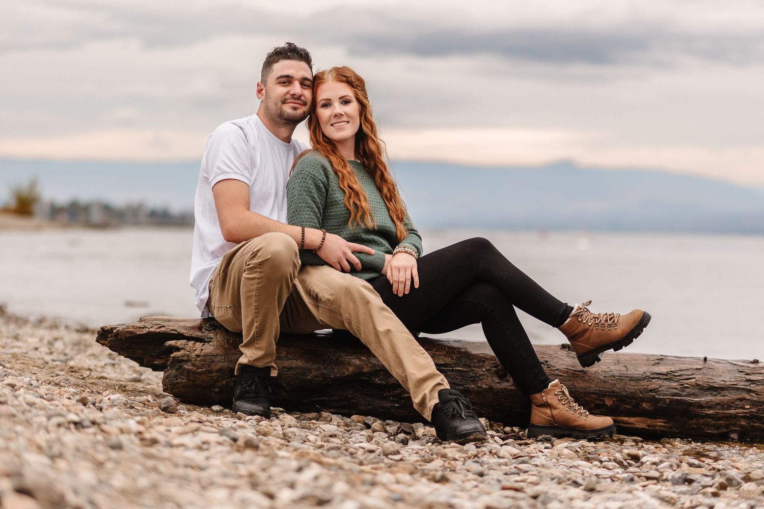 A couple sits together on driftwood at a rocky beach during sunset with mountains in the background.