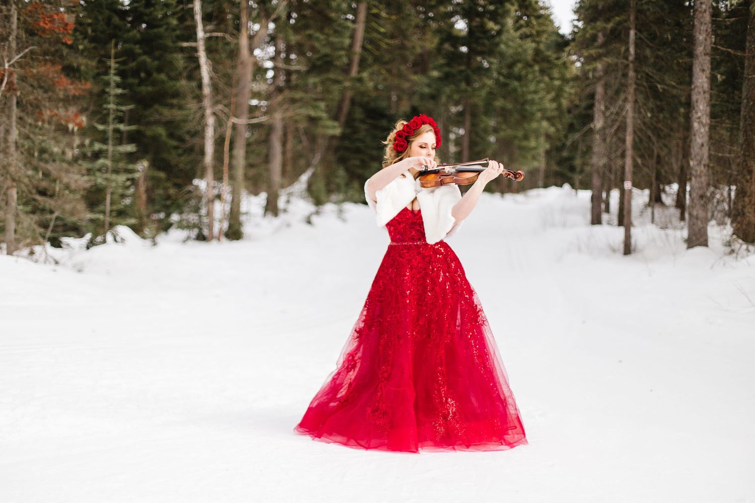 A woman in a flowing red ball gown poses elegantly in a snowy winter forest setting.