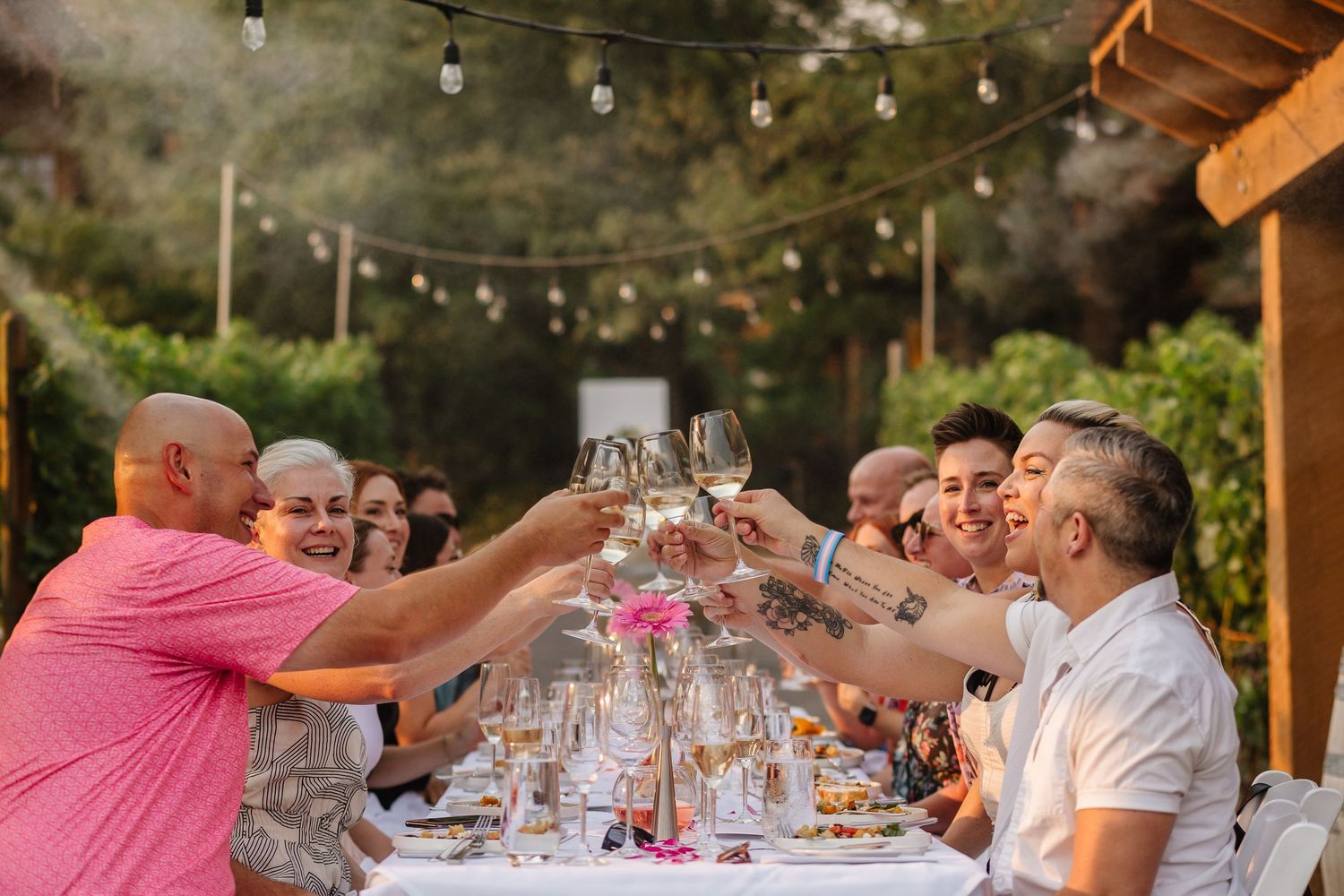 A group of people dining outdoors at a long table under a wooden pergola during a summer gathering.