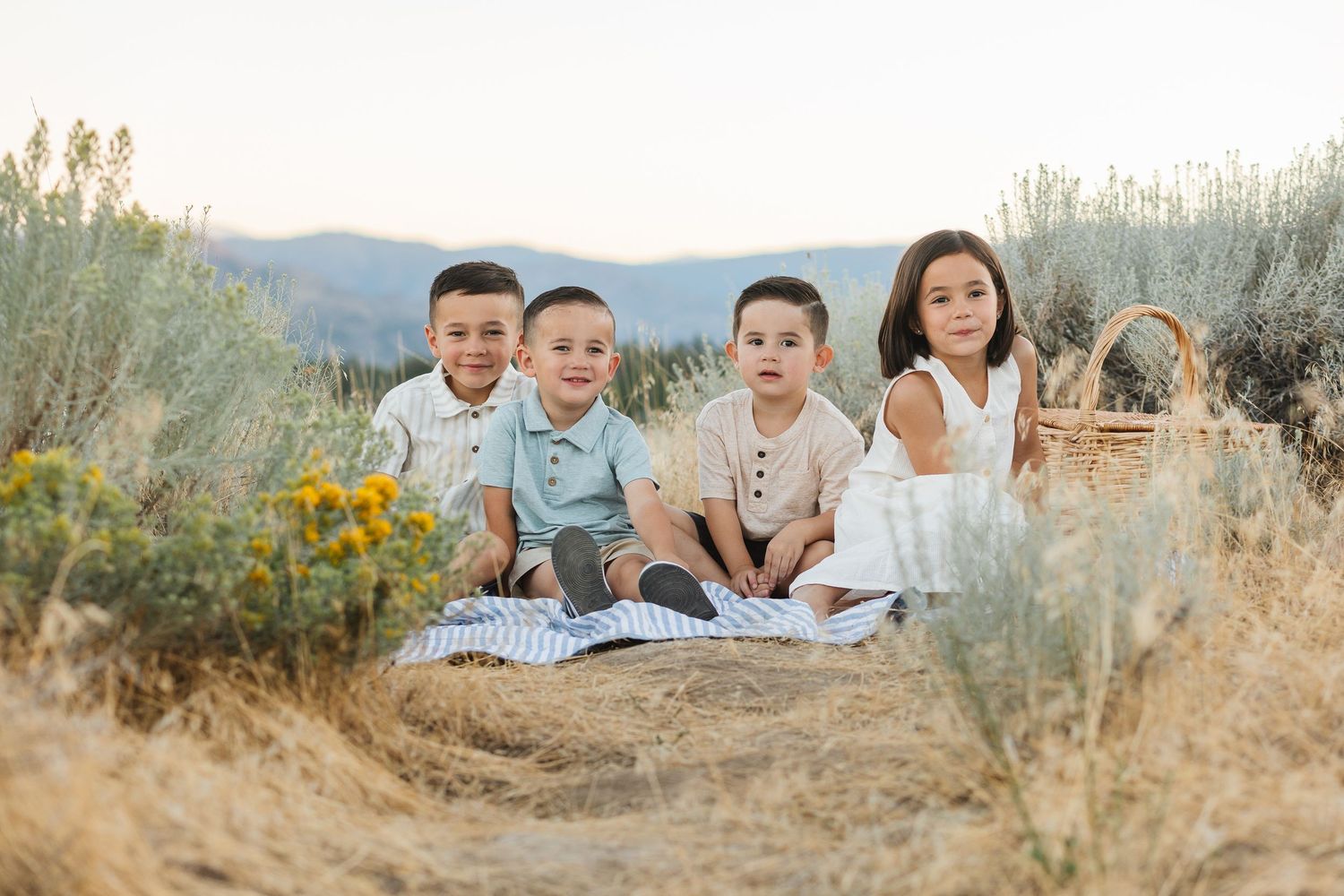 Children sitting on a blanket in a golden field at sunset with mountains in the background.