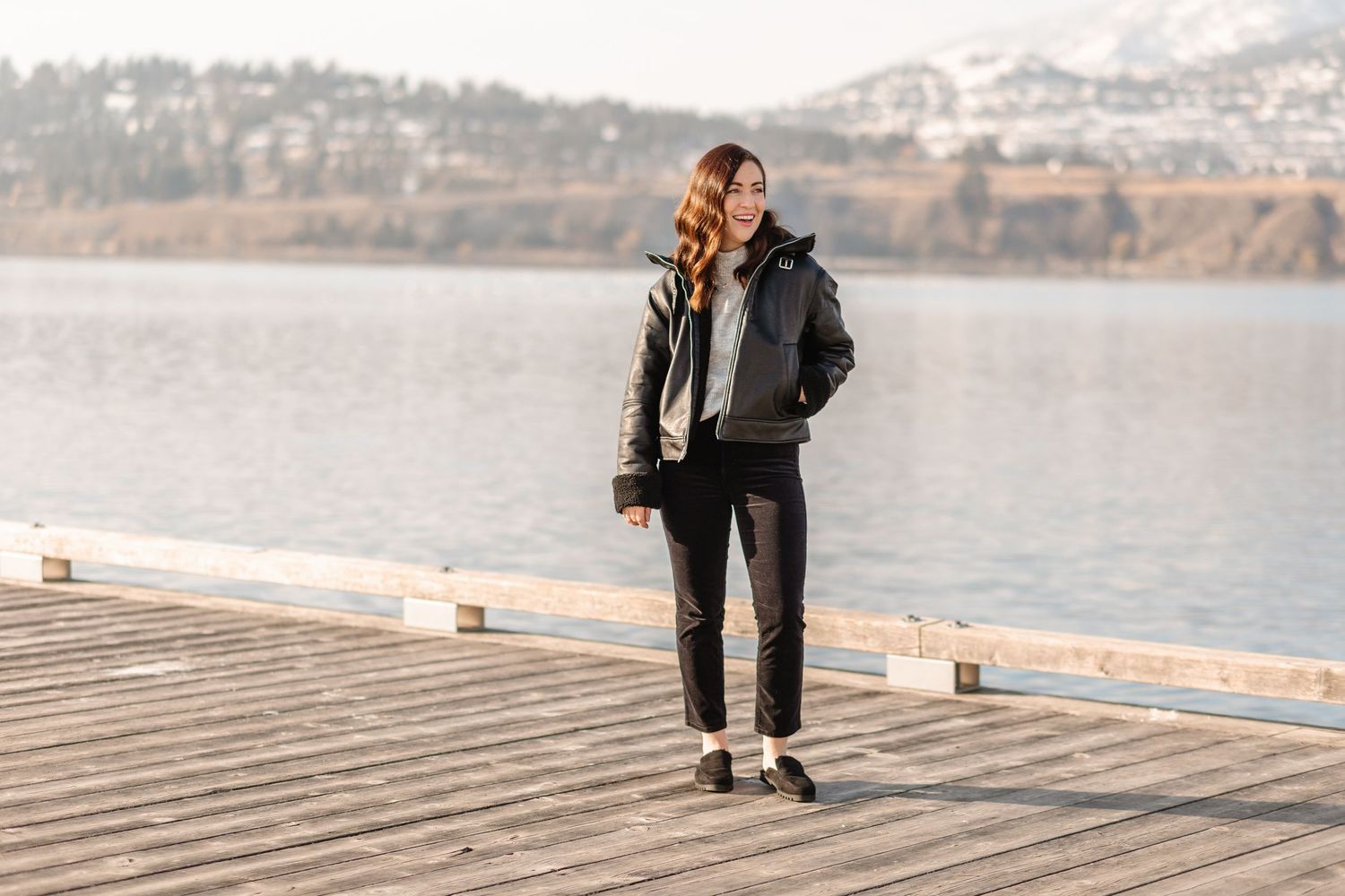 A couple stands together on a wooden dock overlooking a scenic mountain lake on a sunny day.