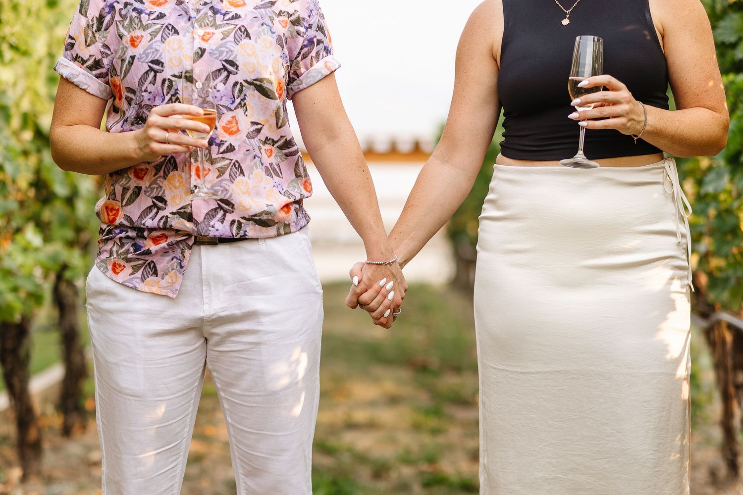 Two people holding hands while carrying wine glasses in a vineyard, wearing floral top and cream skirt.