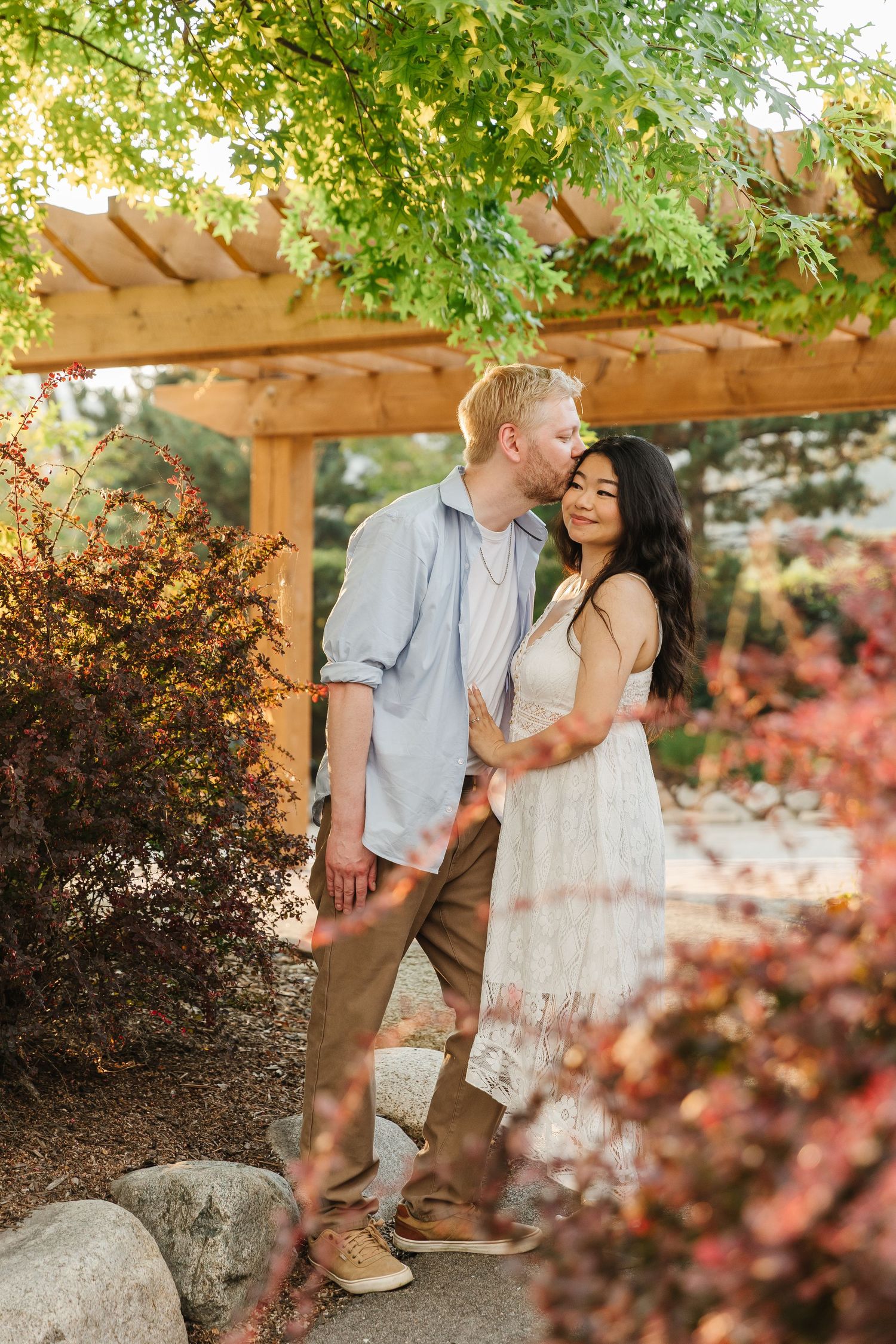 A couple shares an intimate moment under a wooden pergola surrounded by lush greenery and pink flowers.