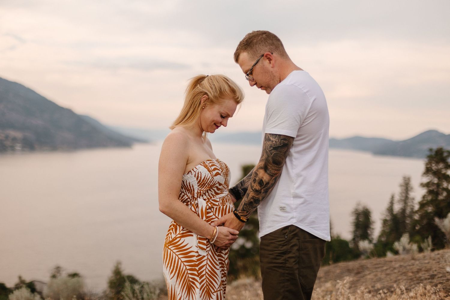 A couple stands on a mountain overlook during sunset, with a serene lake and mountains in the background.