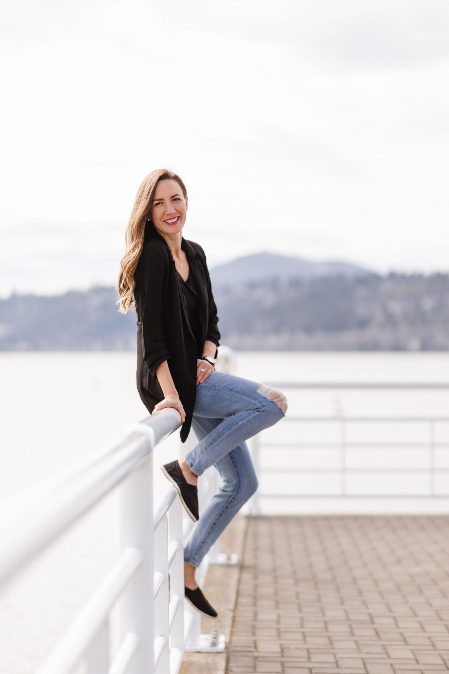 Woman in black top and jeans poses playfully along white railing by waterfront in casual outdoor photoshoot series.