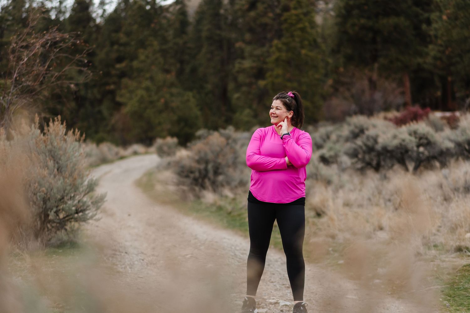 A hiker wearing bright pink walks along a winding desert trail surrounded by sagebrush and pine trees.