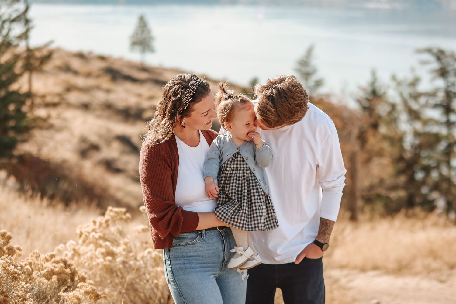 A family shares a tender moment together on a golden hillside with evergreen trees in the background.