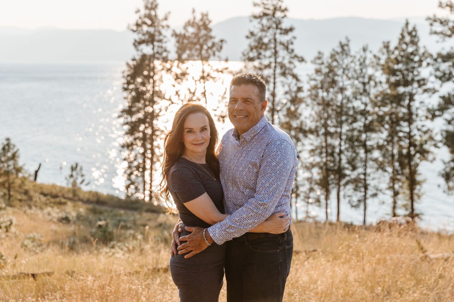 A couple stands together on a grassy hillside overlooking a lake with tall pine trees in the background at sunset.