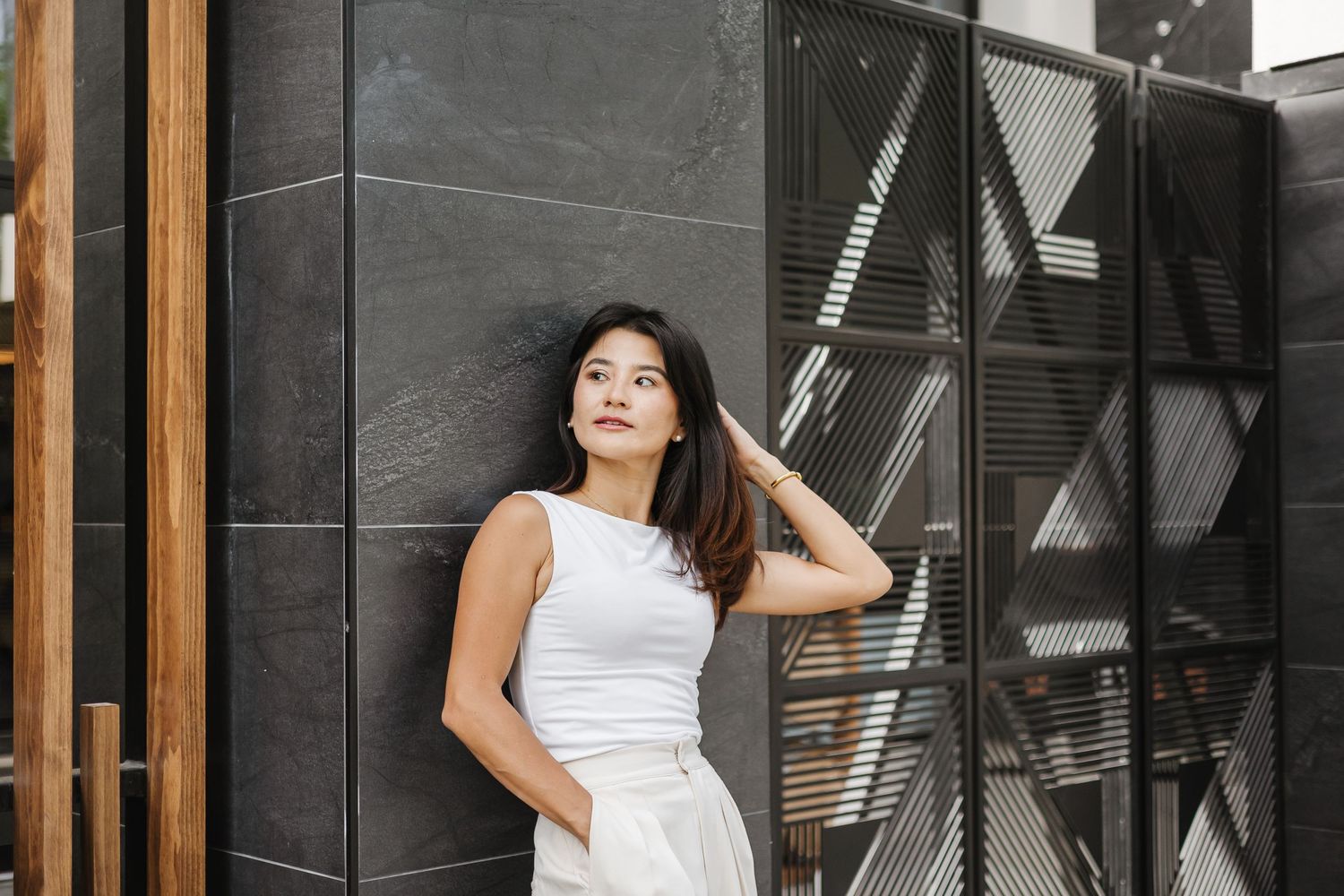 Woman in white sleeveless dress poses against modern building exterior with glass windows and wood accents.