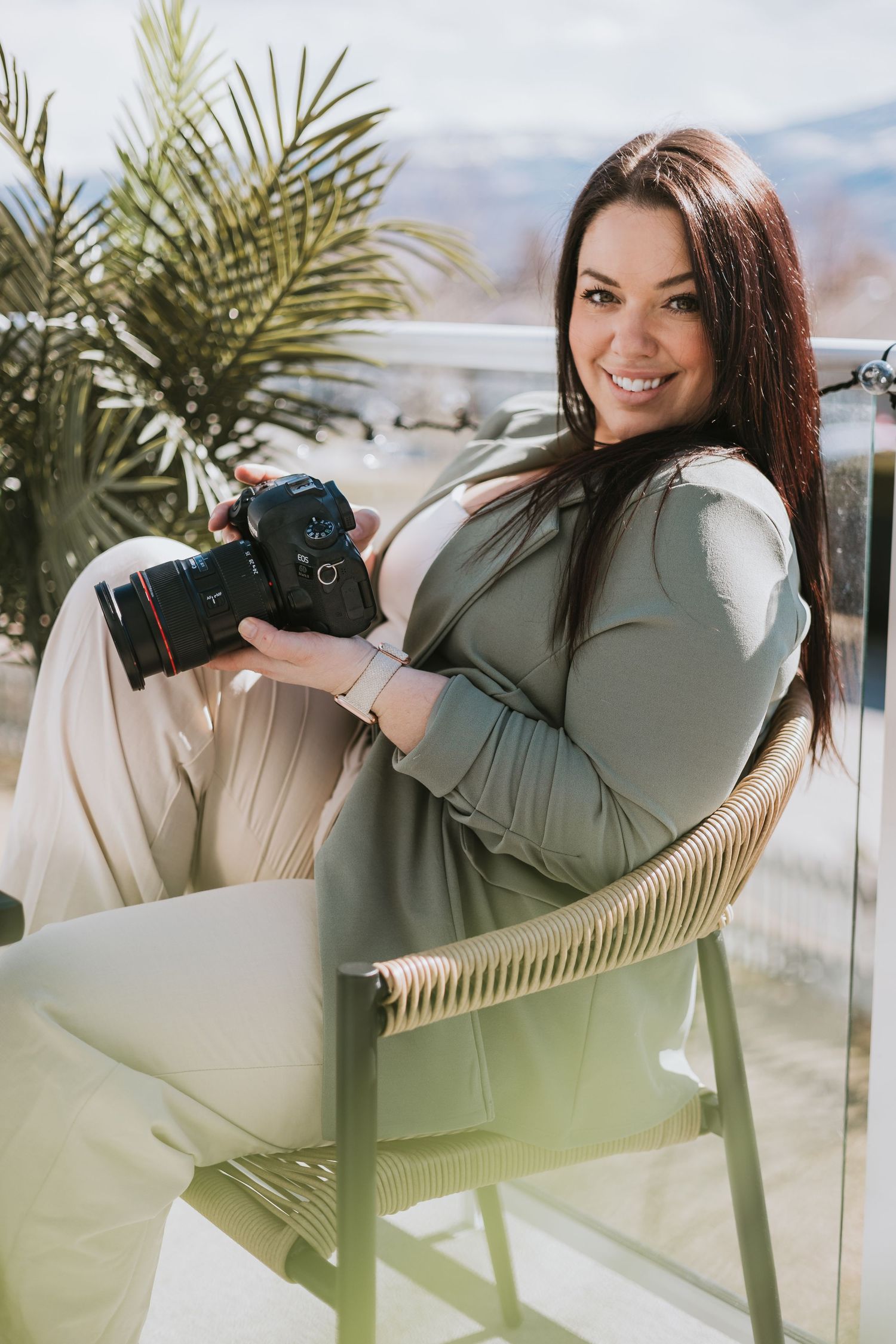 Professional photographer wearing olive green sweater sits in rattan chair with camera near tropical plants in West Kelowna BC