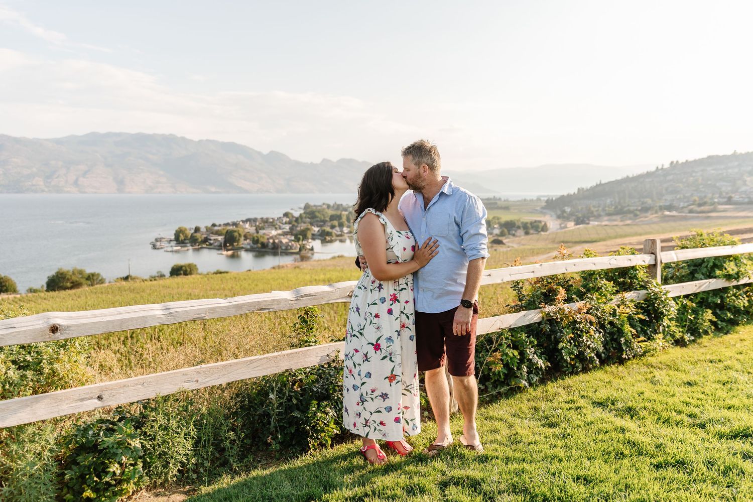 A couple shares a romantic moment on a hillside overlooking a lake and mountains on a sunny afternoon.