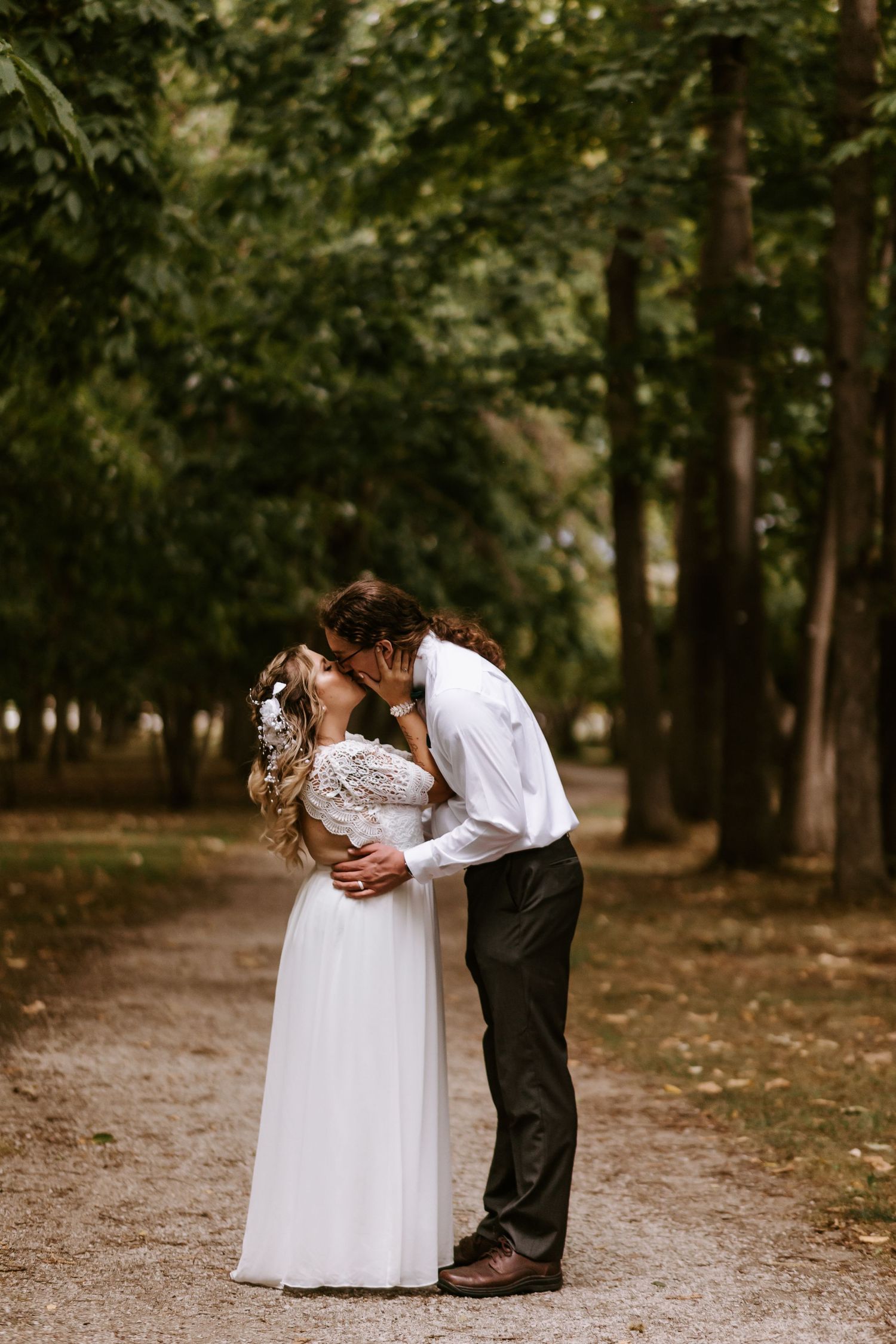 A romantic wedding moment captured in a wooded park setting with a couple embracing on a dirt path.