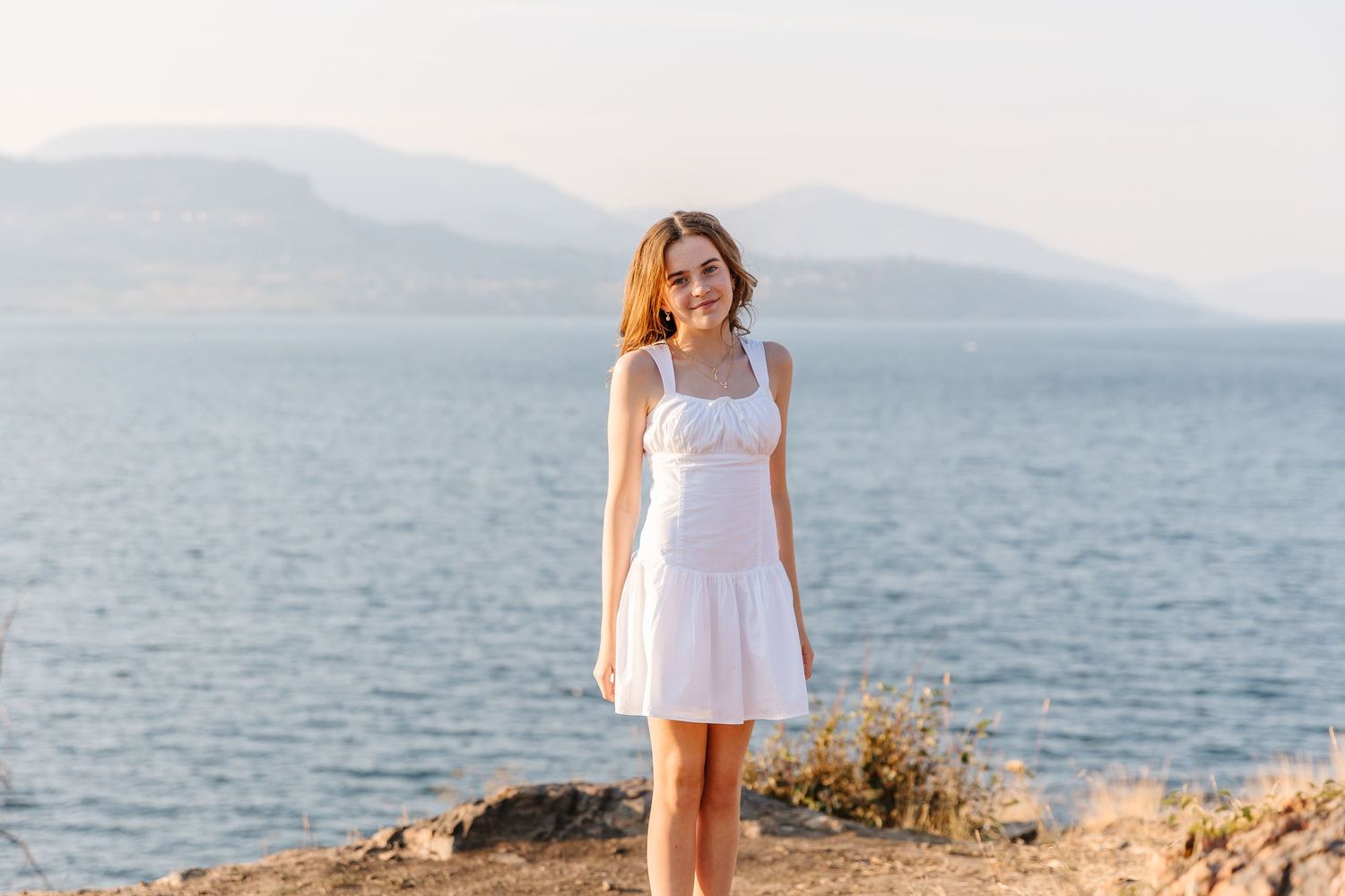 A person in a white sundress stands on rocky coastal terrain with mountains and ocean in the background.