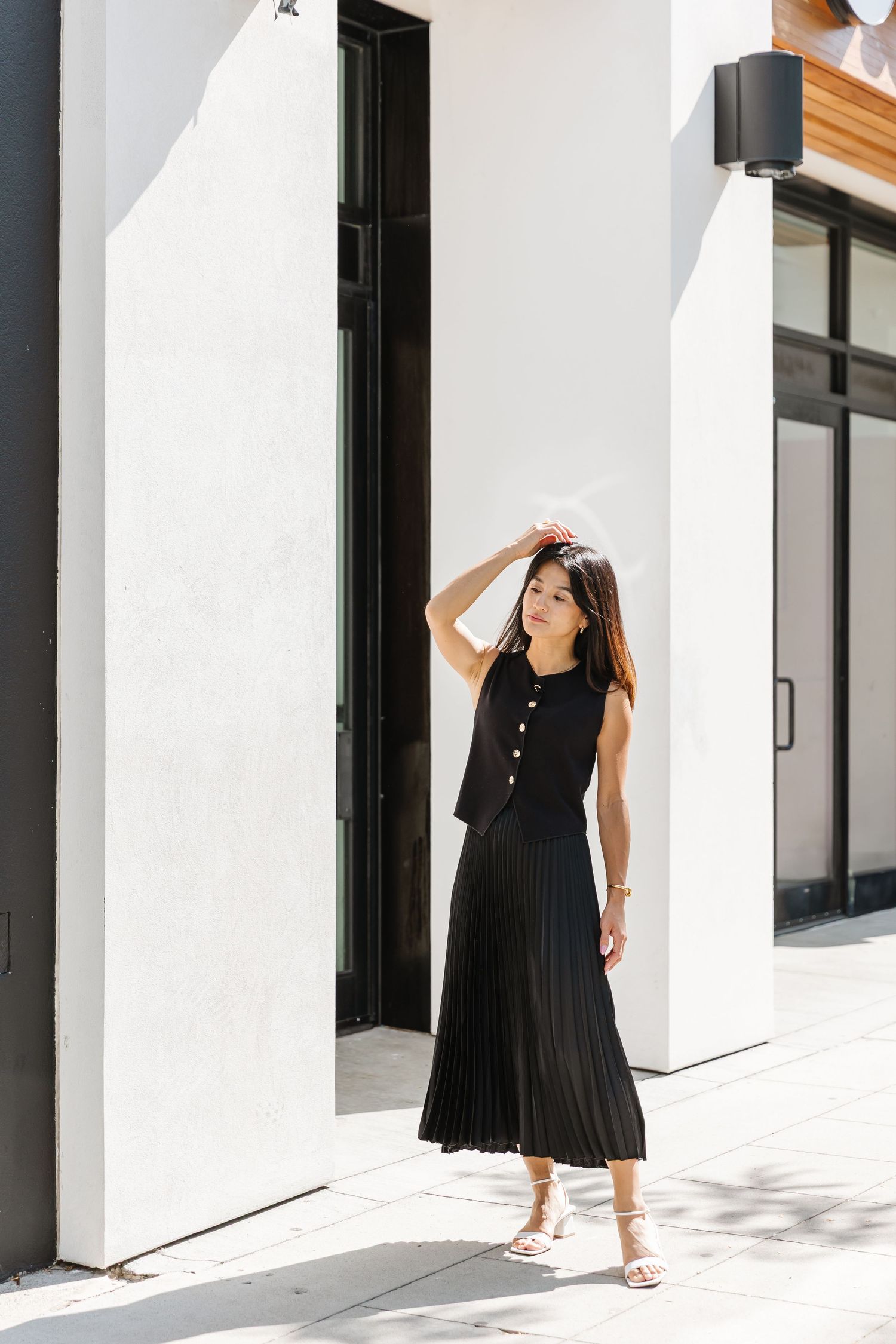 Woman in black sleeveless dress poses gracefully against white architectural backdrop in minimalist outdoor setting.