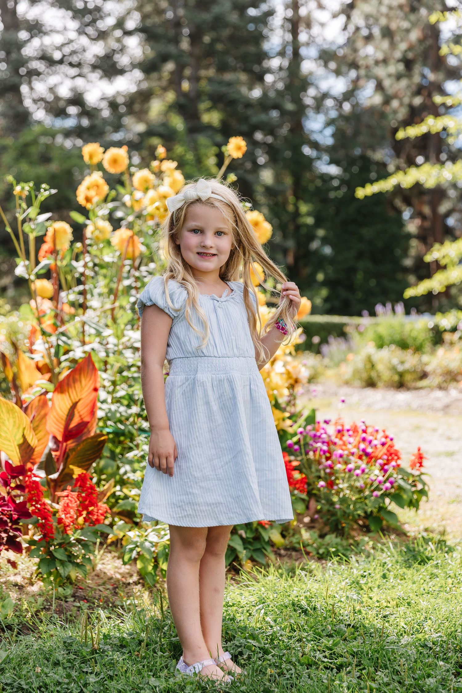 A child in a white dress stands among colorful flower gardens on a sunny summer day.
