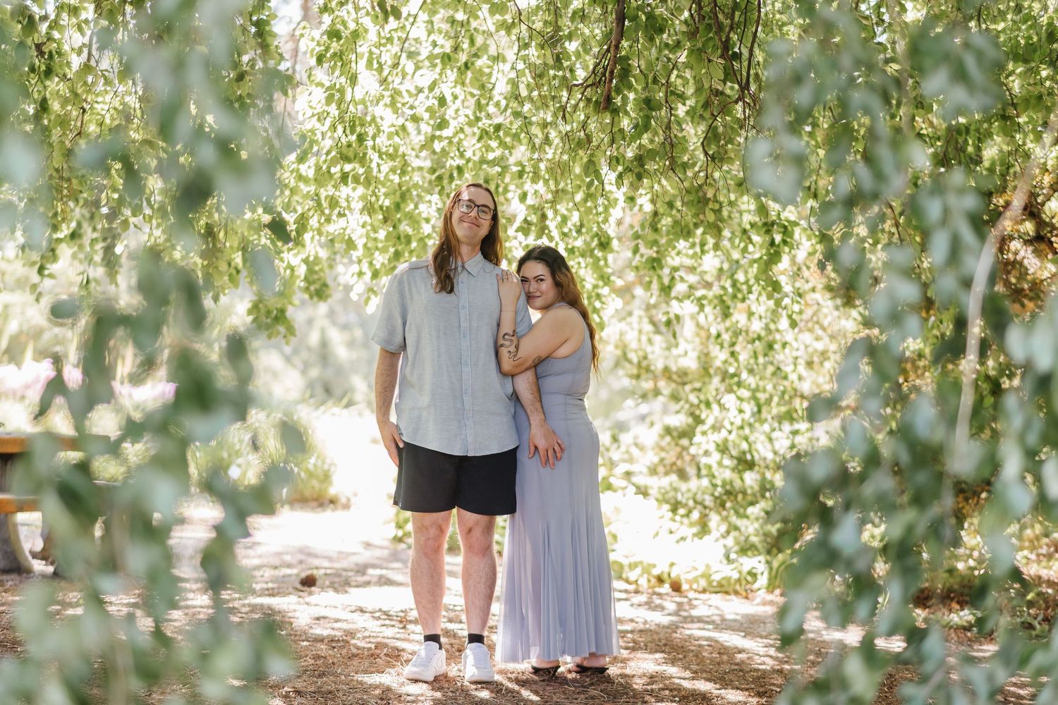 A couple stands together under hanging willow tree branches on a sunny summer day in a garden setting.