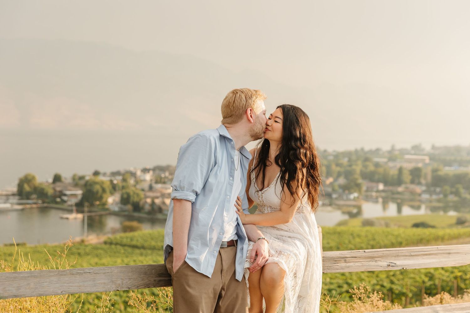 A couple shares a romantic kiss on a hilltop overlooking a scenic waterfront landscape at golden hour.