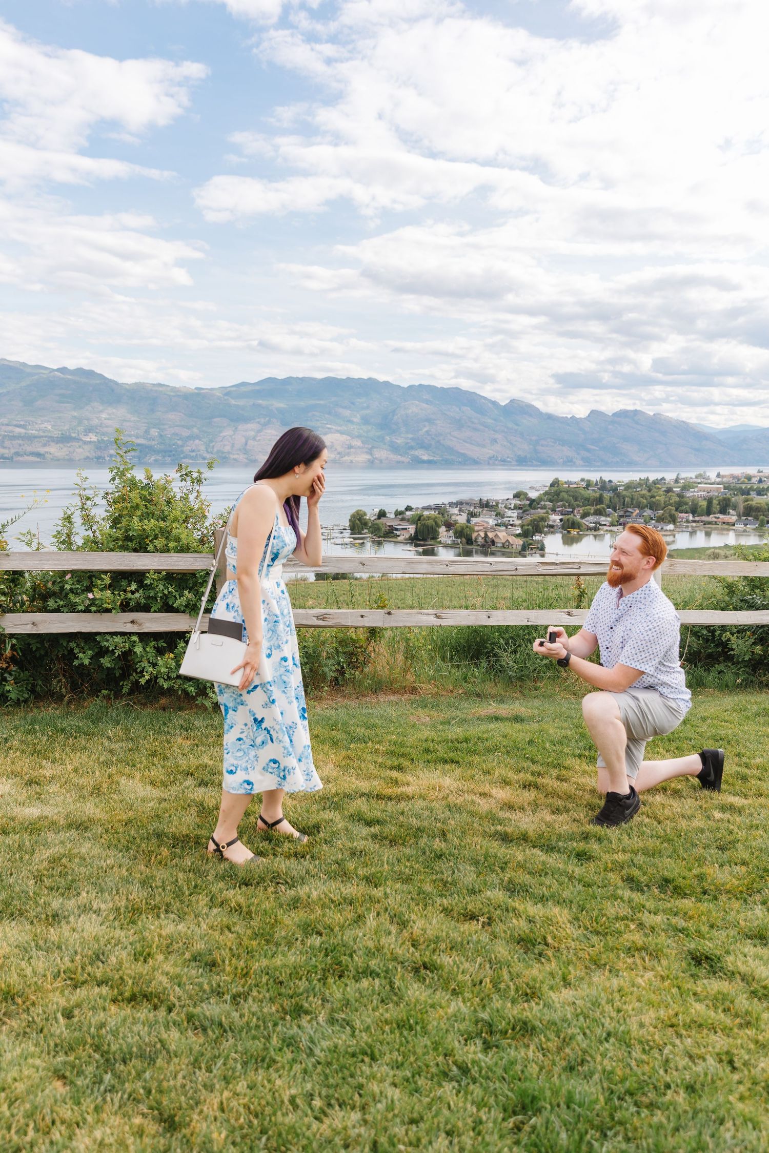 A couple shares a special moment on a grassy hilltop overlooking a scenic mountain and lake vista.