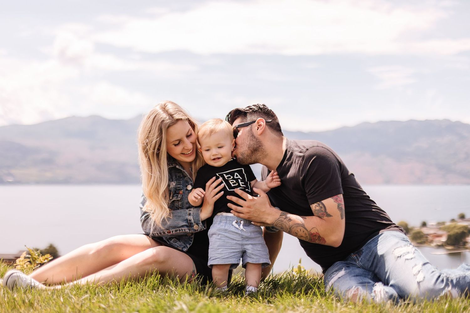 A family in casual clothing shares tender moments together while sitting on a grassy hillside overlooking a scenic lake view.