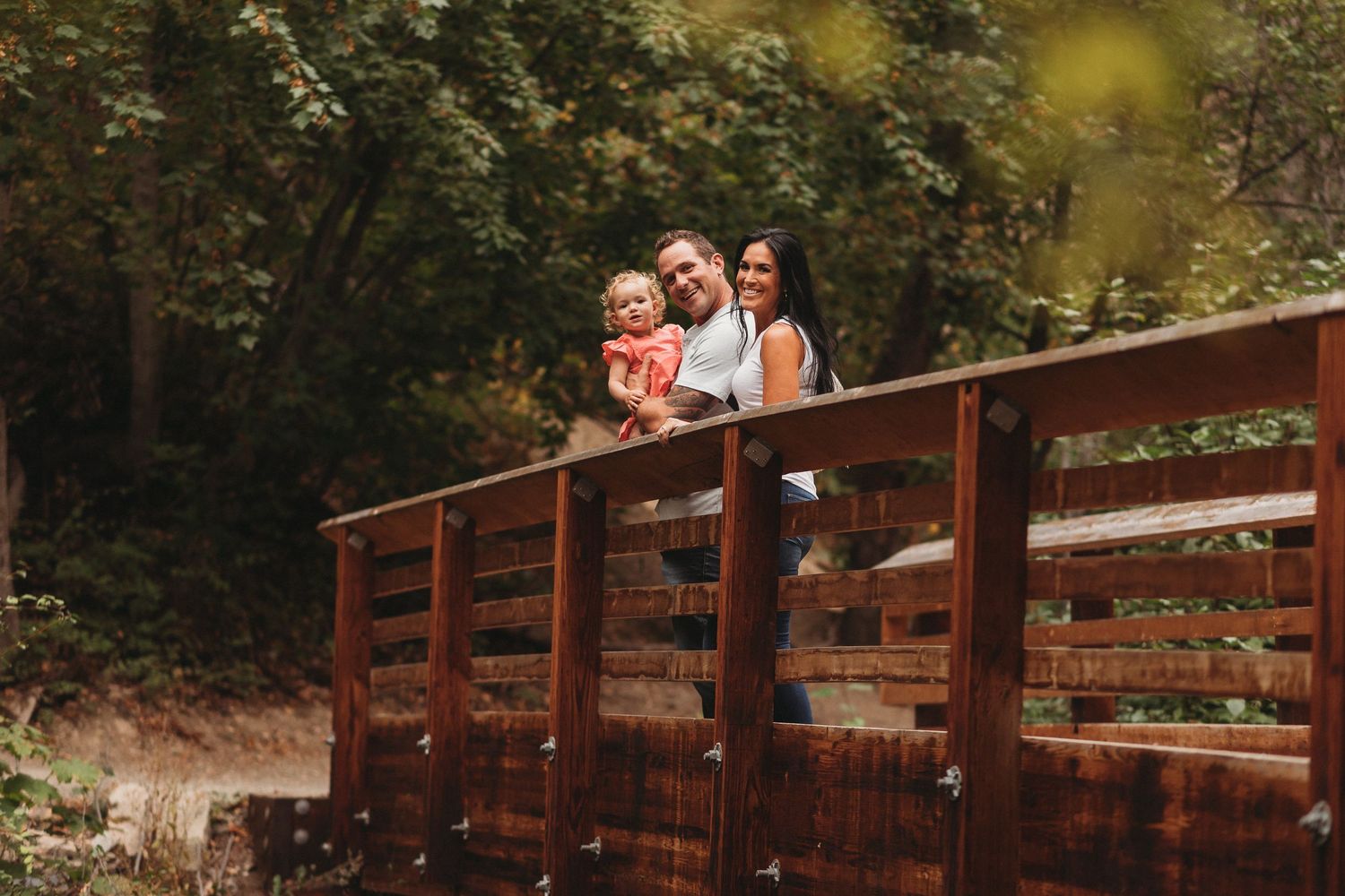 A family stands together on a wooden bridge surrounded by lush greenery on a sunny afternoon.