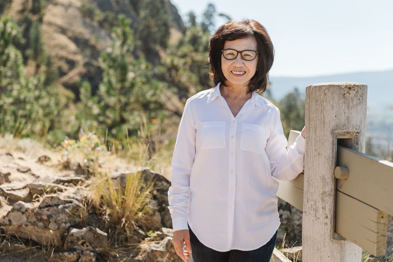 A person in a white shirt stands on a scenic overlook with mountains and natural landscape in the background.