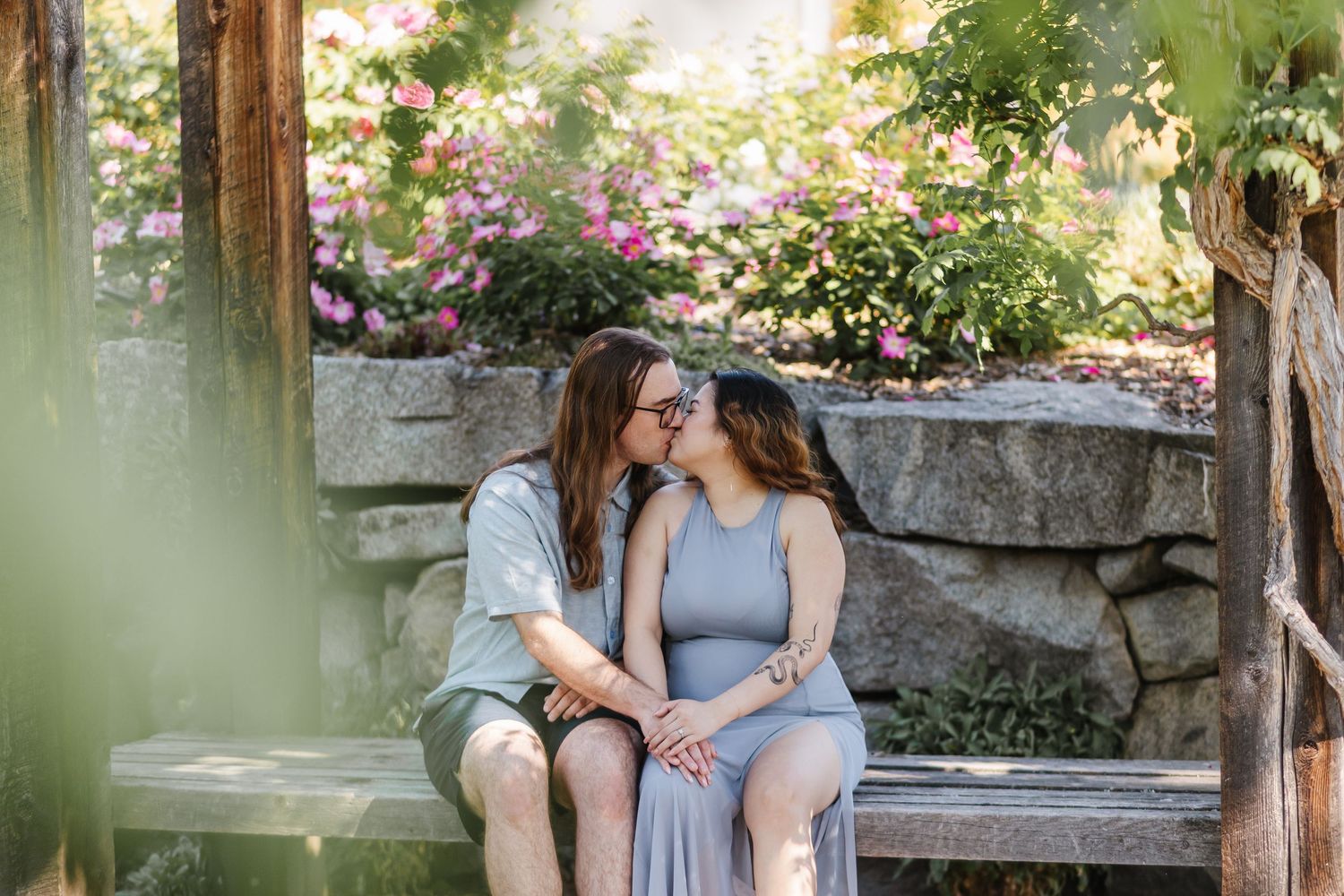 A couple sits together on a stone wall surrounded by lush greenery and flowers in a romantic garden setting.