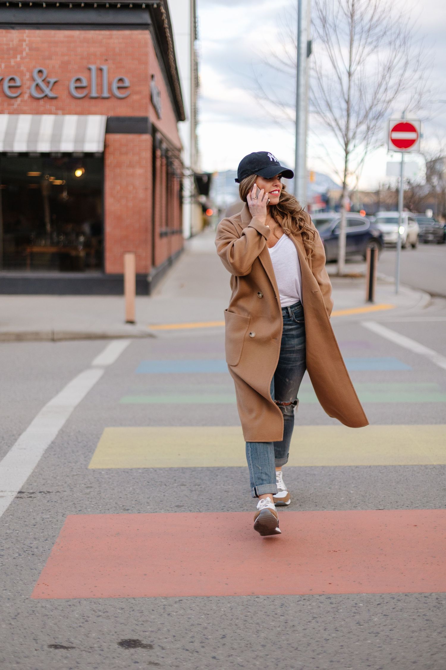Stylish person in camel coat and jeans walking on colorful crosswalk while talking on phone in urban setting.