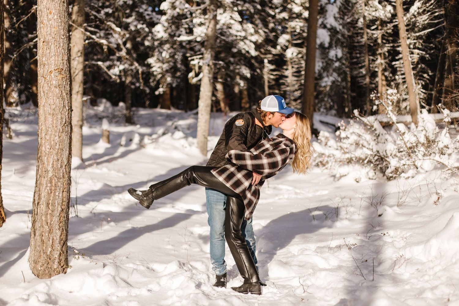 A snowboarder performs a trick jump over a snowy slope between evergreen trees at a winter resort.