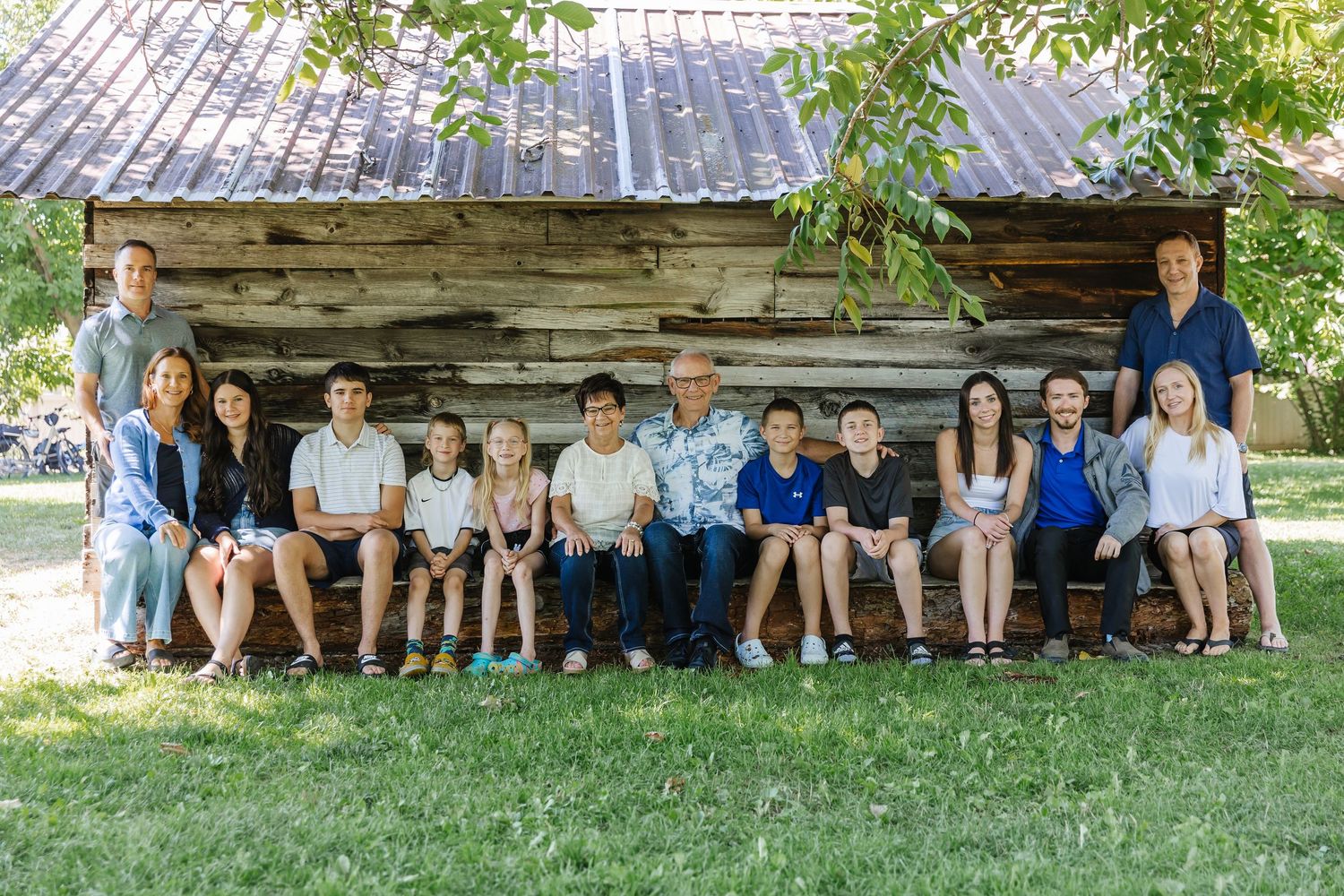 A large group sits together outdoors in front of a rustic wooden cabin on a sunny summer day.