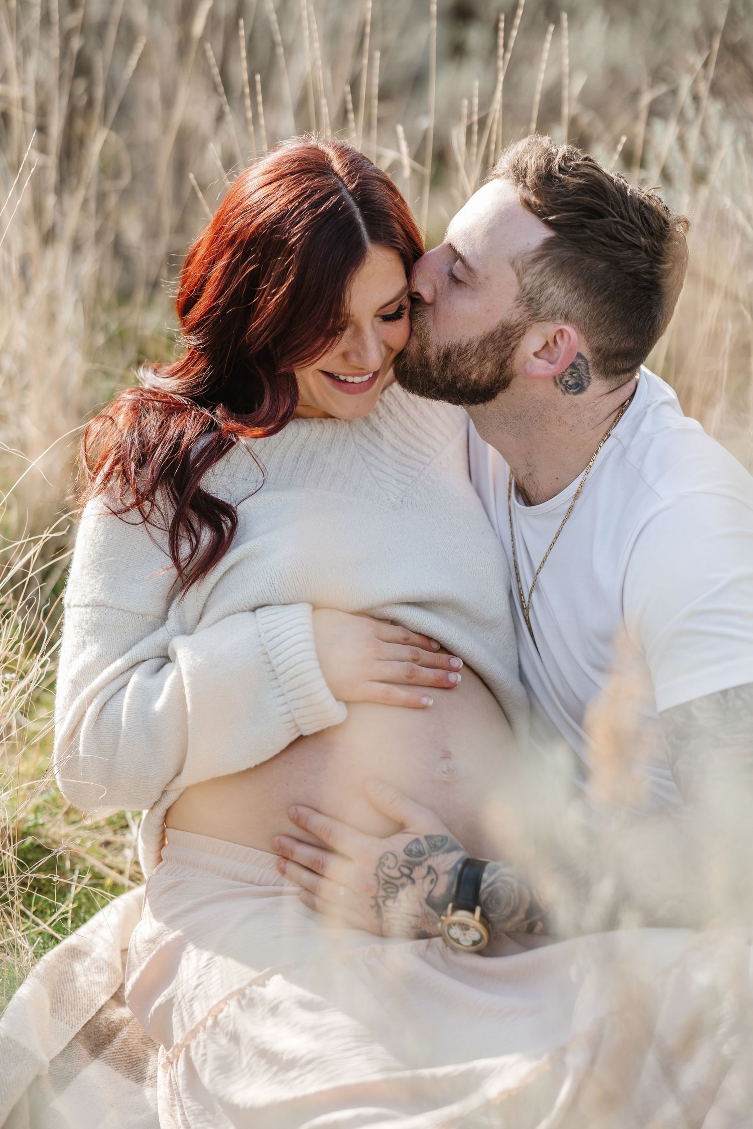 A couple embraces tenderly in a field of tall grass during a maternity photoshoot in soft natural light.