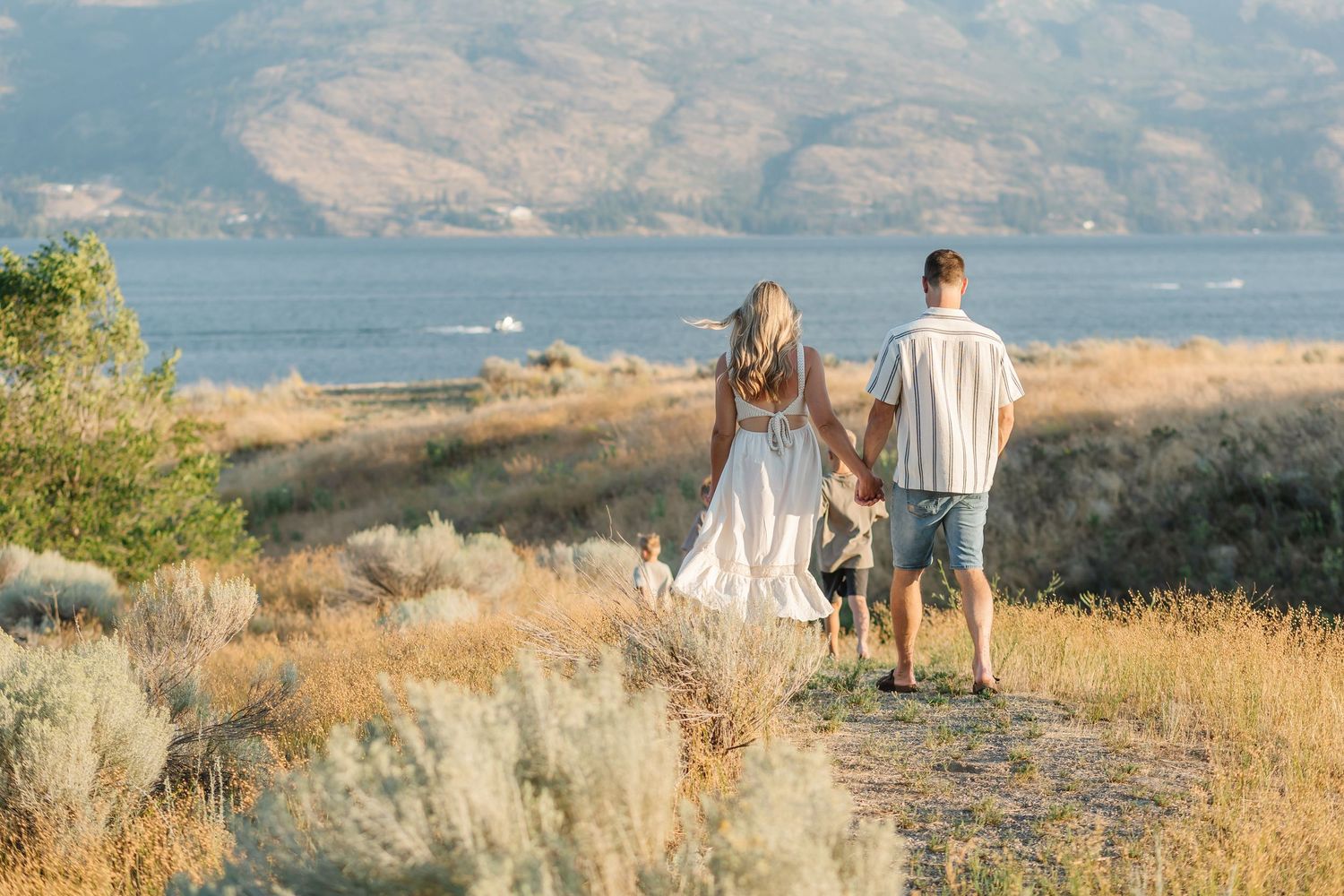 Couple and dog walking on scenic hilltop trail overlooking lake and mountains on sunny summer day.