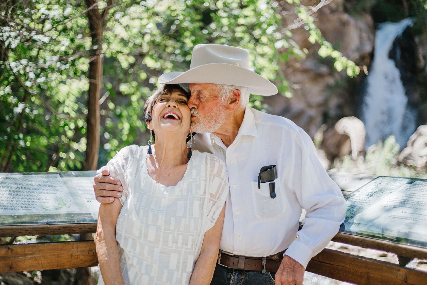 Two people in white outfits sitting at a rustic outdoor table enjoying a conversation in a garden setting.