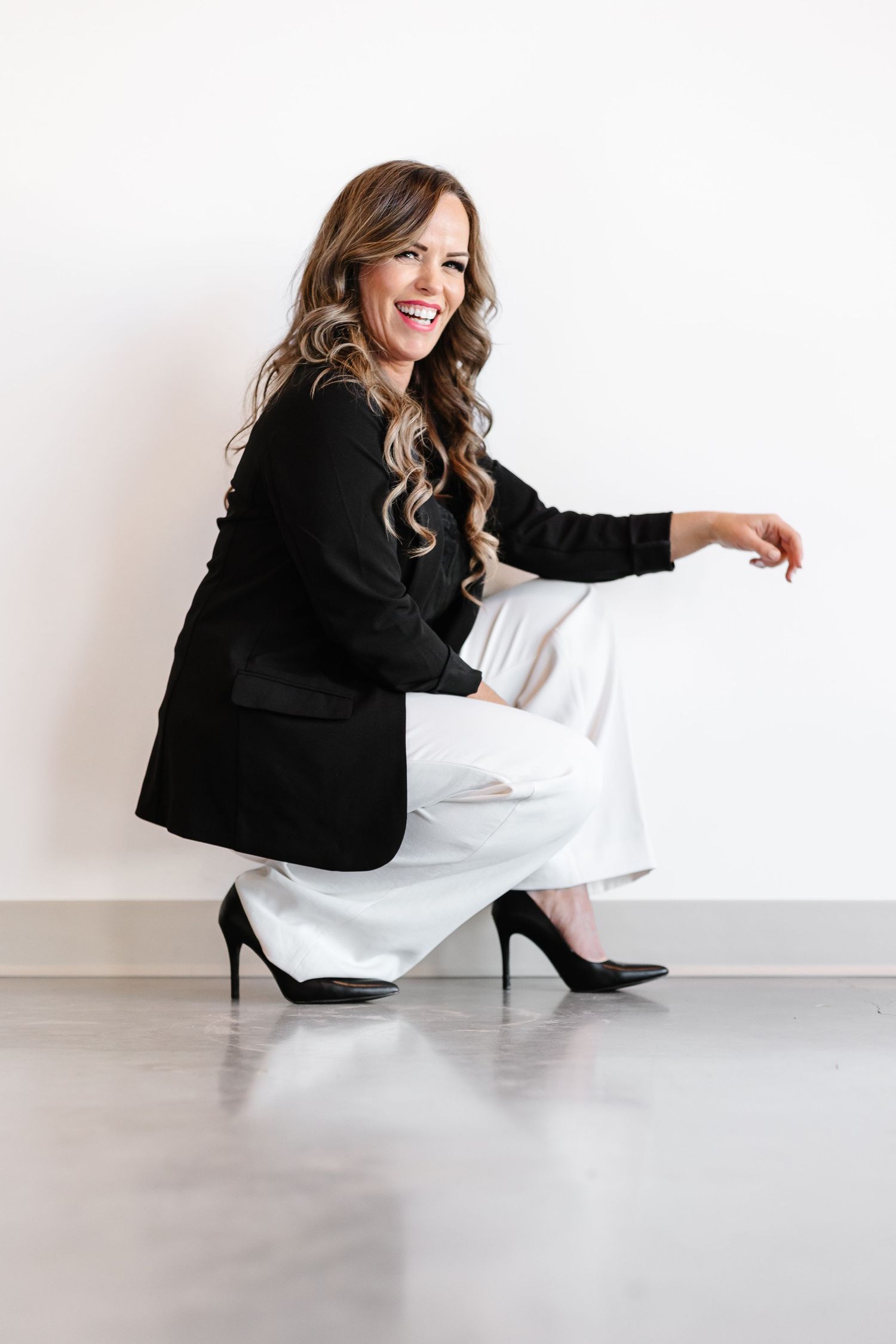 Professional woman in black blazer and white pants crouches elegantly in high heels against white wall.