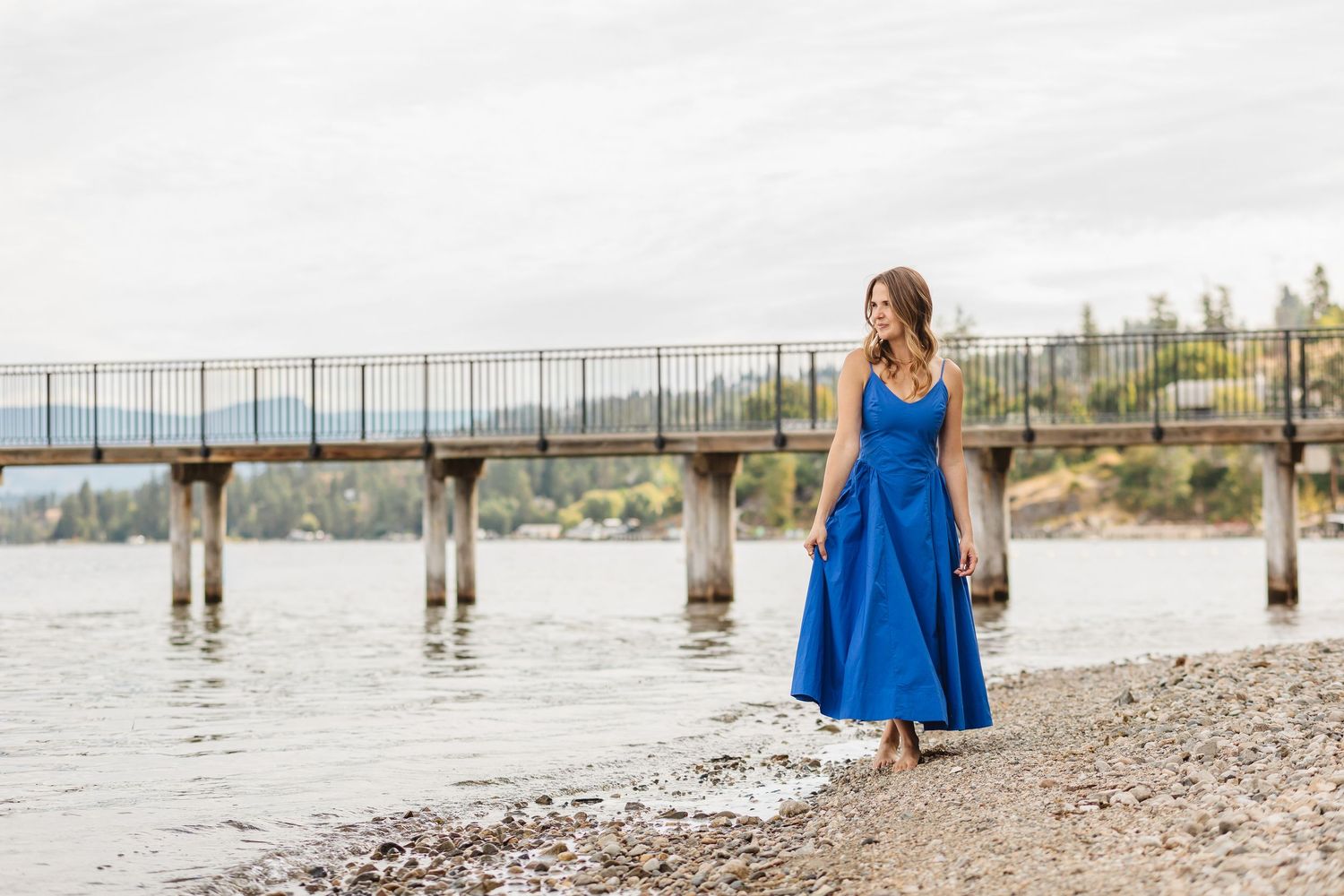 Woman in bright blue dress walks along rocky shoreline near a pier.