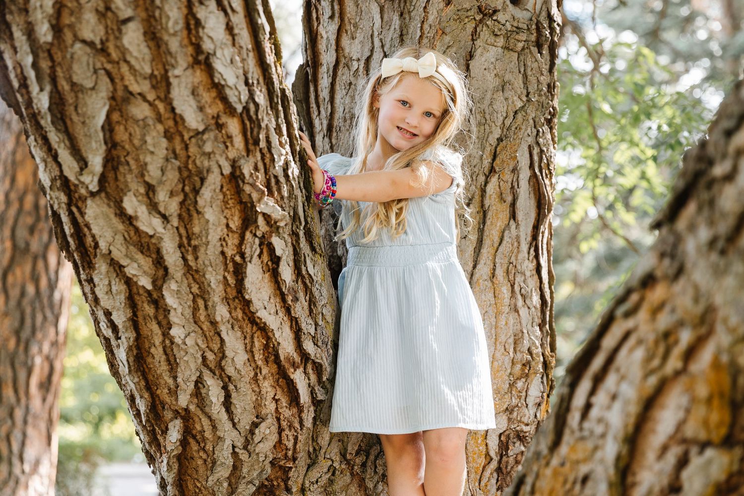 A child in a white dress plays and poses among large tree trunks in a sunny outdoor setting.