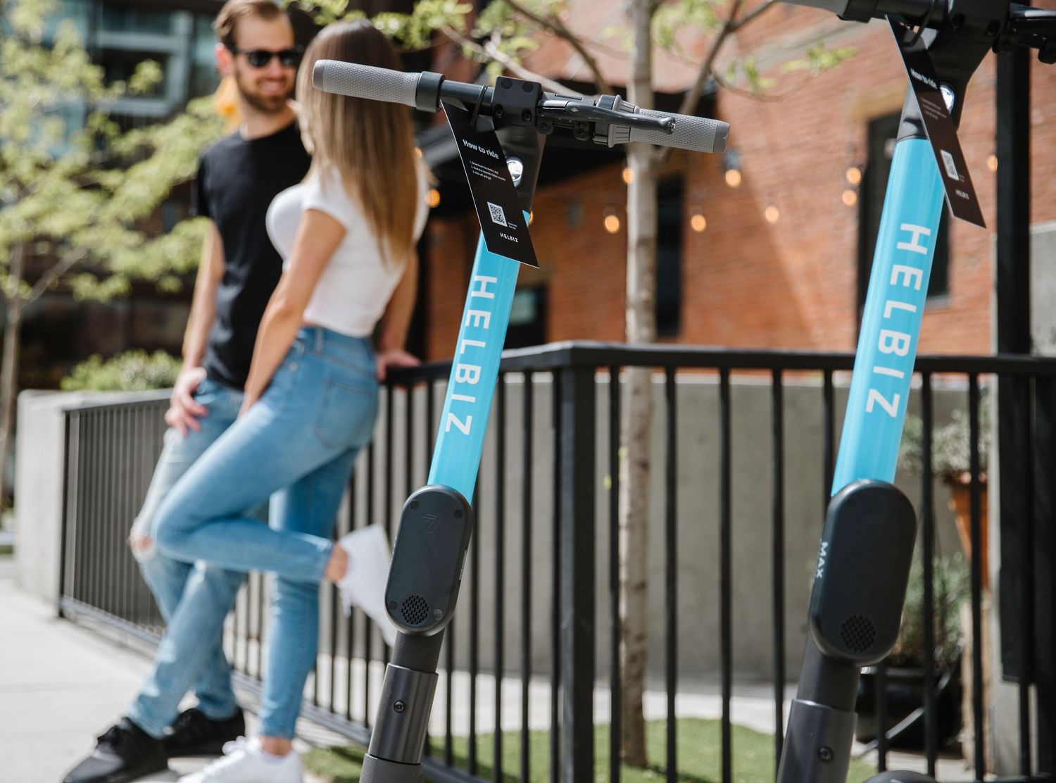 Two blue Helbiz electric scooters parked by a metal railing with people standing nearby.