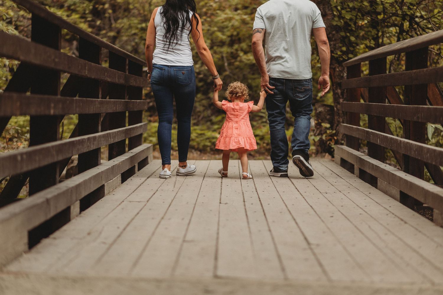 A family walks together holding hands on a wooden boardwalk through a scenic nature area on a sunny day.