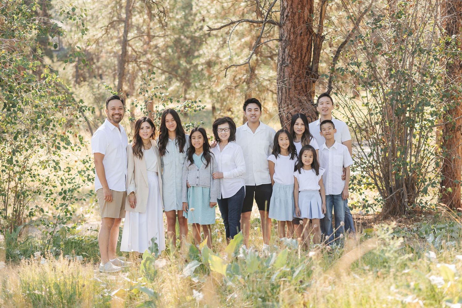 A group in coordinated white outfits poses together outdoors in a sunlit wooded setting for family portraits.