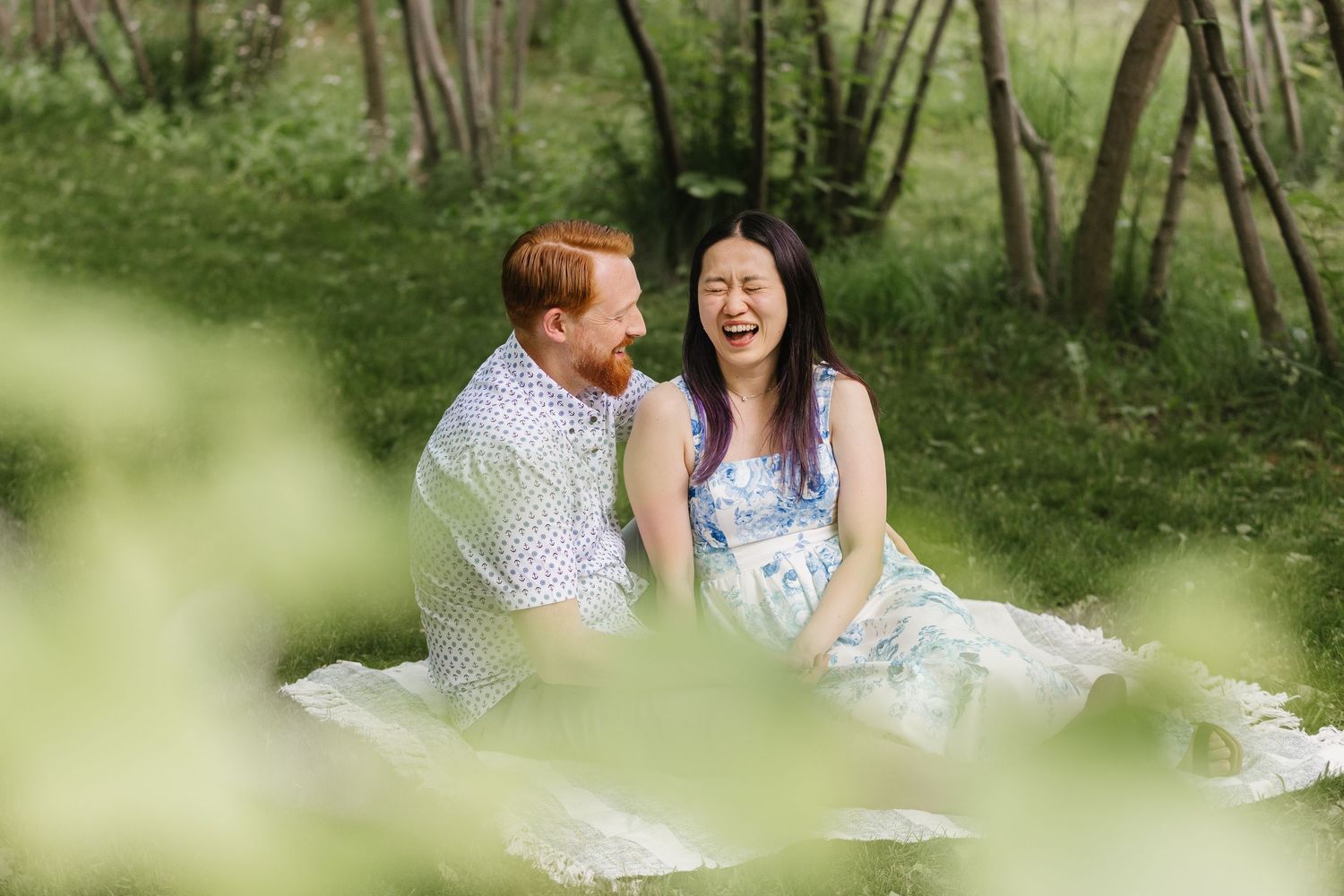 Two people sit together on a blanket having a picnic in a lush green forest setting.