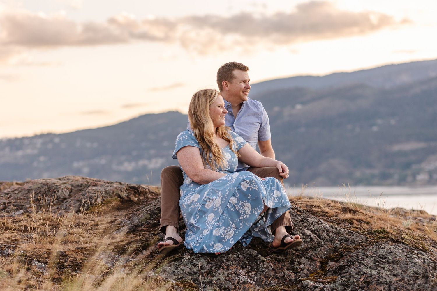 A couple sits on a rocky cliff overlooking a scenic mountain landscape at sunset.